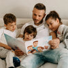 Der Vater hält seine drei Kinder im Arm und liest ihnen ein Buch mit lustigen Geschichten über sie vor.