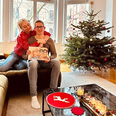 Ein Pärchen lächelt vor einem Weihnachtsbaum mit einem individuell gestalteten Pärchenbuch für ihn.