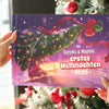 Person hält das Weihnachtsbuch „Sarahs & Mamas – Erstes Weihnachten 2025“ vor einem geschmückten Tannenbaum.