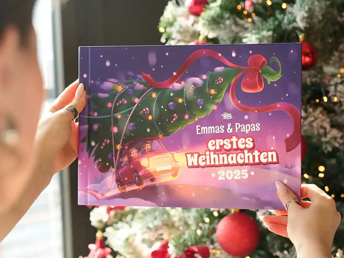 Weihnachtsbuch mit Titel „Emmas & Papas erstes Weihnachten 2025“ vor einem festlich dekorierten Tannenbaum.