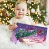 Baby hält das personalisierte Buch „Clara & Paps – Erstes Weihnachten 2025“ vor einem Weihnachtsbaum.